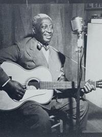 Leadbelly sits on a stool, strumming a guitar, while singing into a standing microphone