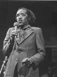 An older Percy wears and dark leisure suit and croons into a mic
