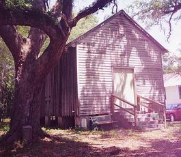 A white clapboard building sits beneath a canopy of trees. Stairs lead to a closed door on the front.