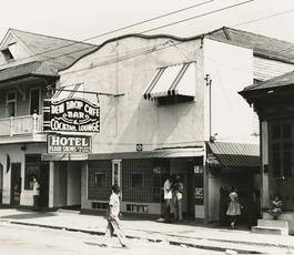 A black and white image of the exterior of the inn, with a sign reading "Dew Drop Inn"; a man crosses the street in front of it