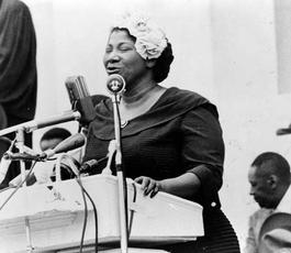 Mahalia Jackson sings from a podium on a stage. She wears a dark dress and has a fancy white hat on