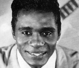 Eddie, in a sharp, light-colored suit and tie, stares into the camera. A guitar is on a backdrop behind him.