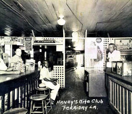 A group of people sit behind a counter top in a nightclub, all turned to face the camera. A younger person sits next to a straw hat. 