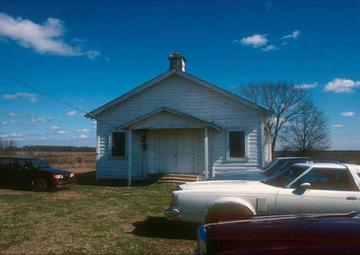 Under blue skies, a white clapboard church sits in a grassy field. 1970s cars are parked outside