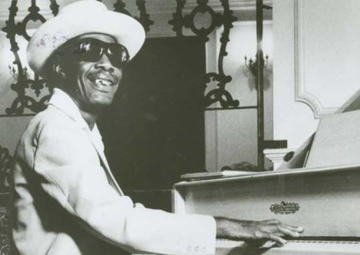 Longhair, in a light-colored suit, straw hat and sunglasses, plays a white piano