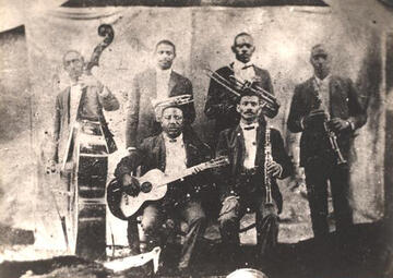 Buddy Bolder and his bandmates; four men stand with their instruments, and two sit; we see a base, guitars, trombones and clarinets