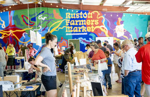 Lively crowd shopping at the busy Ruston Farmers Market in Louisiana, with a colorful mural displaying “Ruston Farmers Market” in the background.