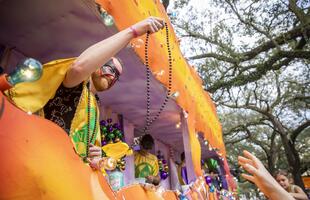 A masked Mardi Gras rider on a brightly decorated float leans out to toss strands of beads to people reaching up from the street below.