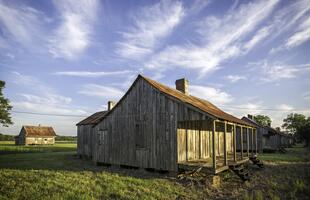 A rustic wooden cabin stands in an open field beneath a sky streaked with wispy clouds.