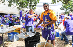 A man decked out in LSU Tigers merchandise stirs a large cast iron pot at a tailgate in Louisiana.