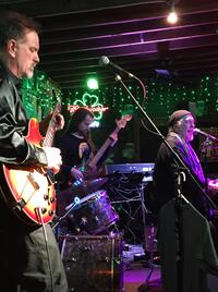 A blues ensemble performs on stage at Enoch's Irish Pub &amp; Grill. Ambient lighting fills a dark stage with shades of violet and green. In the far left side of the frame, a man in a black leather jacket plays a red electric guitar.